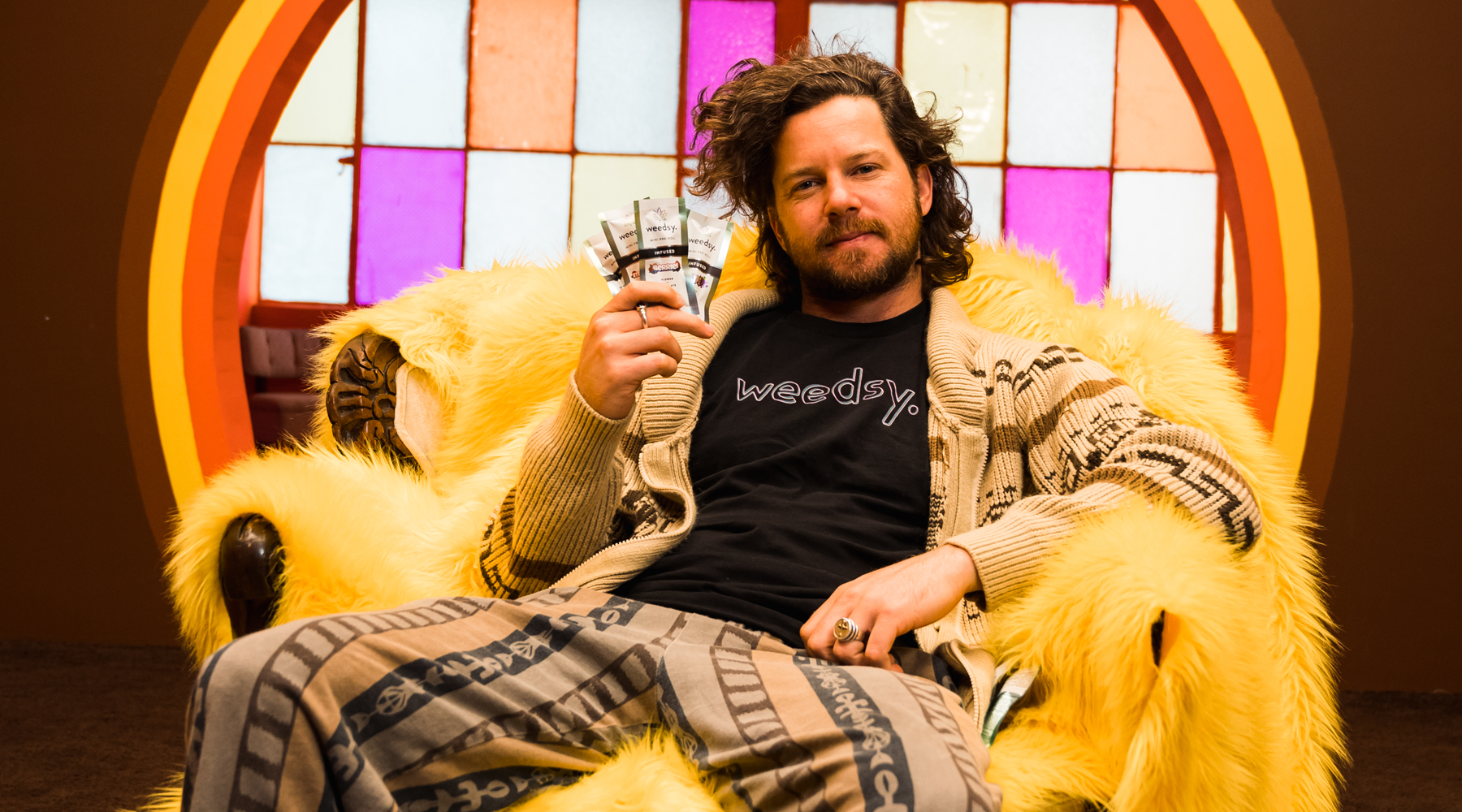 The Dude sitting on a furry chair and holding all six Weedsy Infused mini pre-roll flavors.